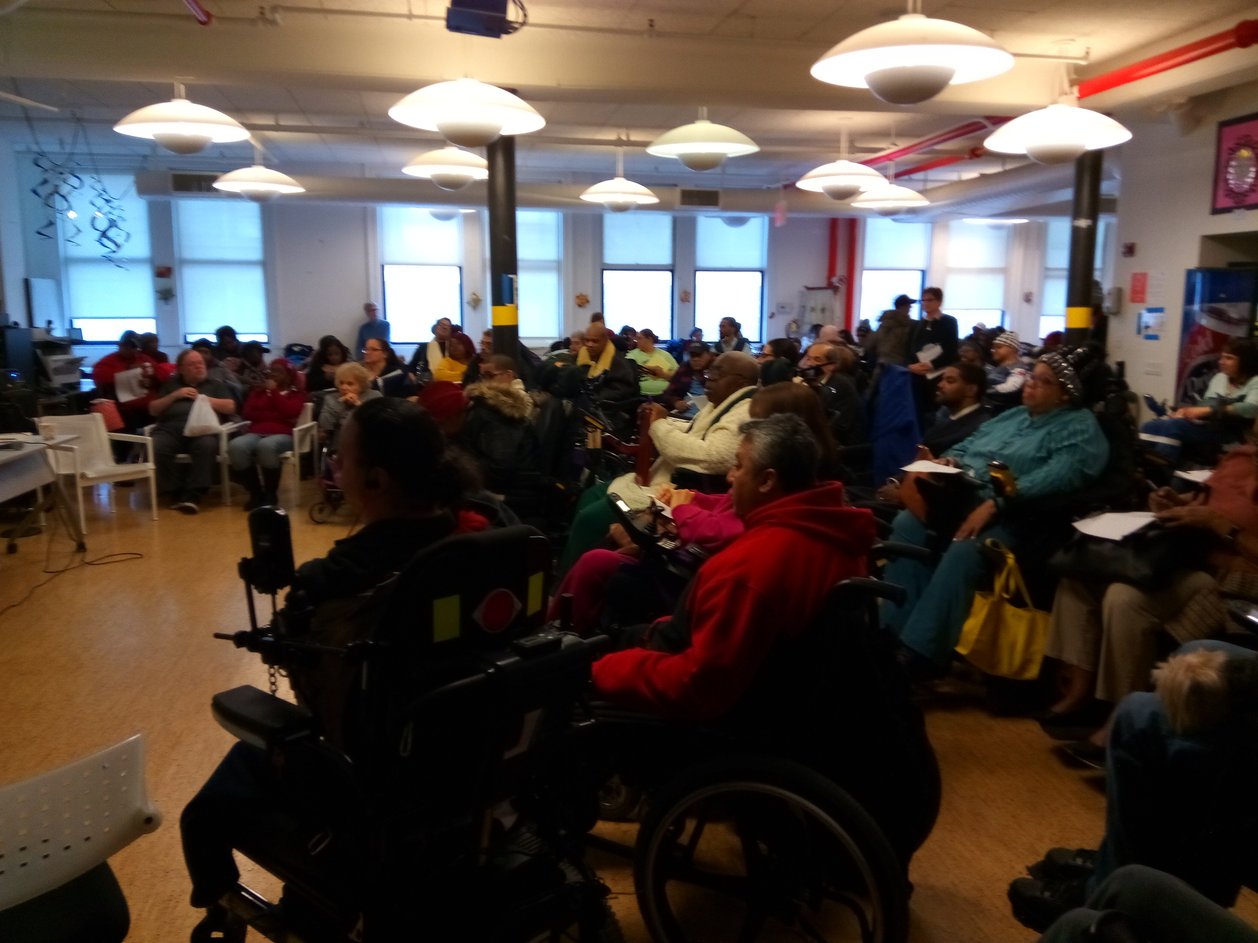 Meeting in a large room with people in chairs
	    	and wheelchairs.
